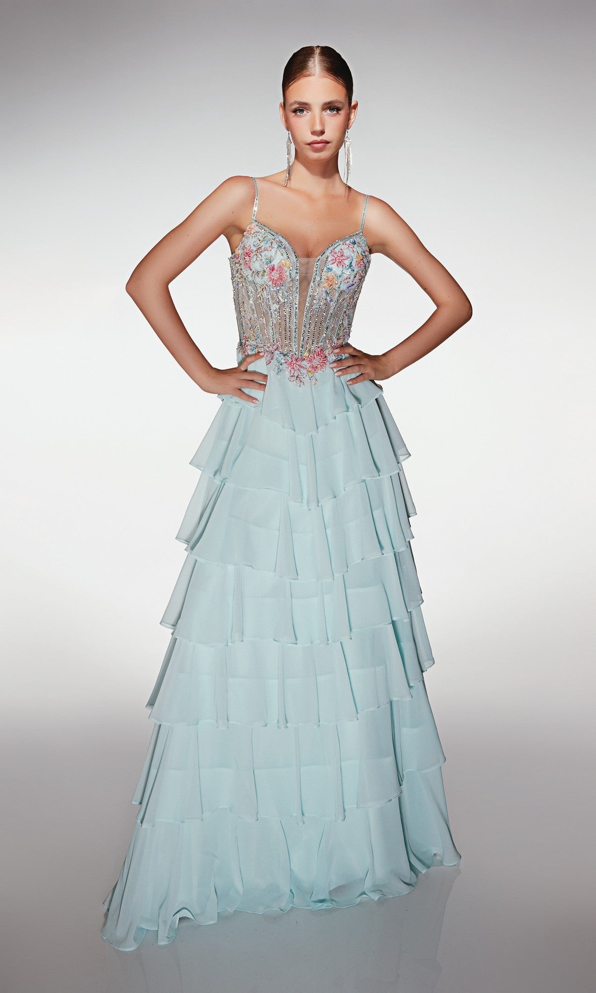 A woman stands with hands on hips, modeling the Alyce 61993 long prom dress in Sea Glass, featuring a chiffon tiered ruffle skirt, thin straps, and a beaded floral corset bodice against a plain white background.